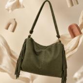 Olive green handbag with a long strap and fringe tassels, suspended in midair against a beige backdrop.
