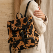Woman in a beige coat wearing a leopard-print backpack with black trim and gold hardware, seen from the side.