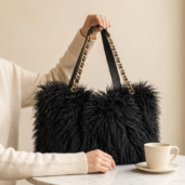 Person in a cream sweater holds a fluffy black handbag with gold chain straps on a marble table, with a coffee cup nearby.