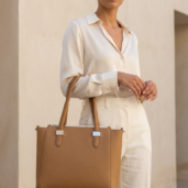 Woman in a cream blouse and matching trousers carries a tan structured leather tote outdoors with silver hardware.