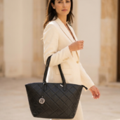 Woman in a beige suit carrying a black quilted tote with a round pendant charm hanging from the strap.