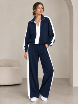 Woman in a navy tracksuit with white side stripes, standing with one hand in her pocket in a minimal studio setting.