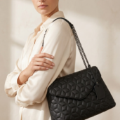 Woman in a light blouse carries a black quilted shoulder bag with a chain strap.