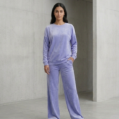Woman standing in a lavender velour loungewear set against a concrete wall, hands in pockets.
