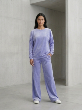 Woman standing in a lavender velour loungewear set against a concrete wall, hands in pockets.