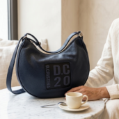 Navy blue leather handbag with a D.C.20 logo patch on a marble table; a person in beige sits nearby with a cup of coffee.