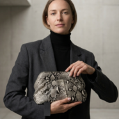 Woman in a dark blazer holds a snakeskin-patterned clutch in front of a neutral concrete backdrop.