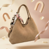 Beige tote bag with a patterned scarf tied to the handle, suspended in soft pastel backdrop with rose petals nearby.