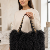 Woman in a beige sweater holding a large black fluffy tote with gold chain straps.