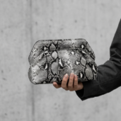 Person's hand holds a snakeskin-pattern clutch bag against a concrete wall.