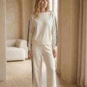 Woman wearing a cream tracksuit with black side stripes standing in a softly lit room by sheer curtains.