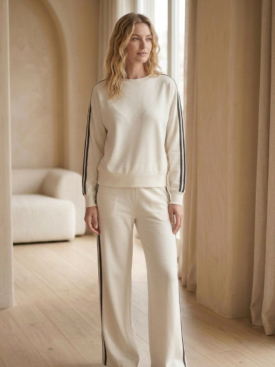 aNeh-UrYaA4cyOYotOykEd - Merlin Luxury Woman wearing a cream tracksuit with black side stripes standing in a softly lit room by sheer curtains.