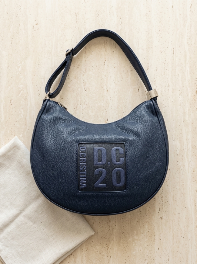 Navy leather shoulder bag with a rounded crescent shape and a square DC20 patch on the front.