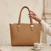 Tanned leather handbag on a marble table with a hand holding its strap, a small espresso cup nearby in a cozy cafe setting.