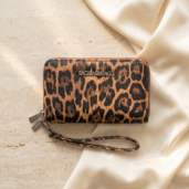 Leopard print zip wallet with a matching wrist strap resting on a light beige fabric and stone surface.