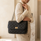 Woman in a cream blouse and beige pants carries a black quilted leather handbag with a gold chain strap over her shoulder.