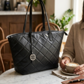 Black quilted tote bag on a marble table in a cafe, with a person holding a cup of coffee nearby.