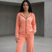 Woman standing in a peach velour tracksuit with zip hoodie and wide-leg pants in a concrete setting.