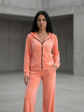Woman standing in a peach velour tracksuit with zip hoodie and wide-leg pants in a concrete setting.