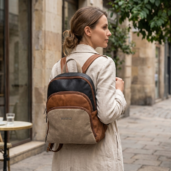Woman in a beige trench coat walks along a cobblestone street with a two-tone brown and gray backpack on her back, seen from the side.