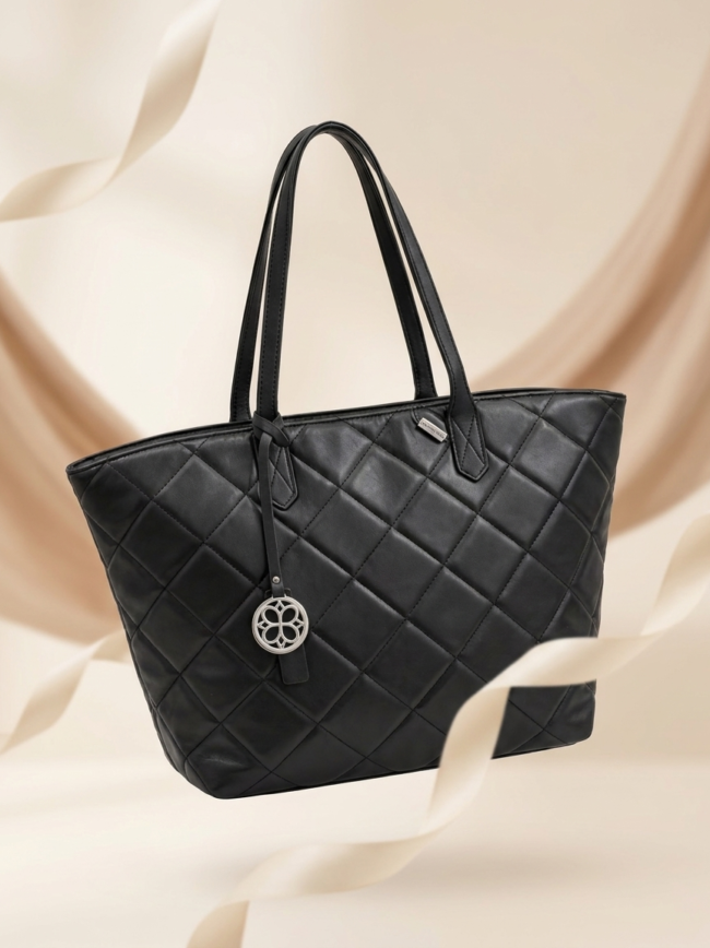 Black quilted tote bag with double handles and a silver circular charm, against a beige fabric backdrop.