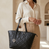 Woman in a cream blouse and beige pants holding a large black quilted tote bag with a circular metal charm pendant.