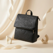 Black textured leather backpack with flap and gold zipper, displayed against a cream background with decorative ribbons (product photo).
