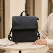 Black textured leather backpack with top handle and adjustable straps resting on a marble round table; a person in a beige sweater sits beside it.