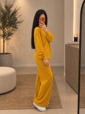 Woman in a mustard-yellow lounge set (sweatshirt and wide-leg pants) taking a mirror selfie with an orange phone in a neutral, modern room with plants and a rug.