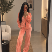 Woman in a peach velour tracksuit taking a mirror selfie in a modern living space, with white sneakers and long dark hair.