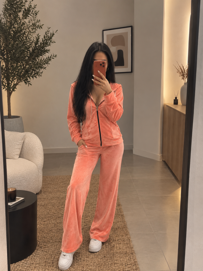 fec52d3c-aa19-4cf3-ad3d-1f1fa98d39b4 1 Woman in a peach velour tracksuit taking a mirror selfie in a modern living space, with white sneakers and long dark hair.