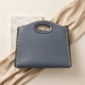 Blue leather handbag with prominent whipstitch chain edging and oval cutout handle resting on beige fabric and marble tile backdrop.