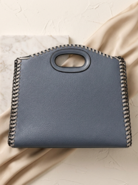 Blue leather handbag with prominent whipstitch chain edging and oval cutout handle resting on beige fabric and marble tile backdrop.