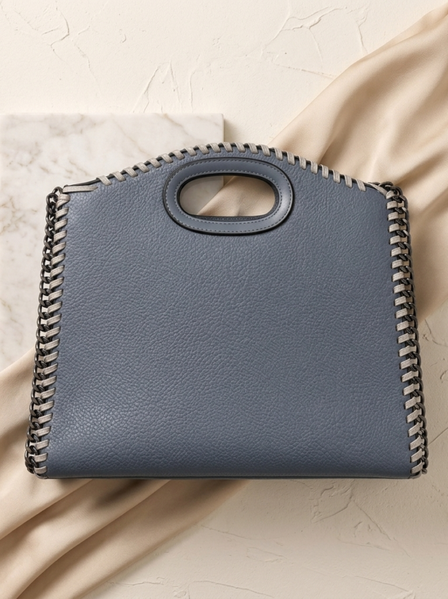 grey blue 2 Blue leather handbag with prominent whipstitch chain edging and oval cutout handle resting on beige fabric and marble tile backdrop.