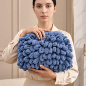 Woman wearing a beige blouse holds a large blue chunky-knit handbag with pom-pom textures in front of her.