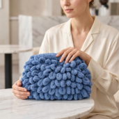 Woman in a beige blouse holds a large blue chunky-knit handbag with looped pom-poms.