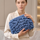 Woman in a light beige outfit holds a large blue chunky-knit handbag with a soft, textured surface across her chest.