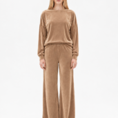 Full-body shot of a model wearing a taupe velour loungewear set with an oversized long-sleeve top and wide-leg pants, paired with white sneakers, facing the camera.