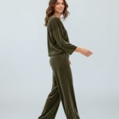 Woman in olive velvet loungewear jumpsuit walking, white sneakers, against a pale backdrop in a studio setting.