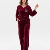 Woman in burgundy velvet loungewear set with a V-neck top and wide-leg pants, logo on the chest, walking forward in white sneakers against a white background.