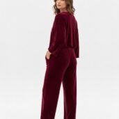 Woman wearing burgundy velour lounge set (top and wide-leg pants) standing in profile with hands in pockets, white sneakers, studio background.