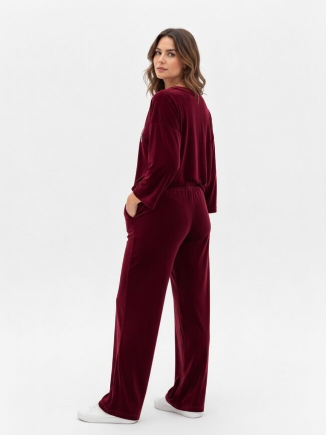 Woman wearing burgundy velour lounge set (top and wide-leg pants) standing in profile with hands in pockets, white sneakers, studio background.