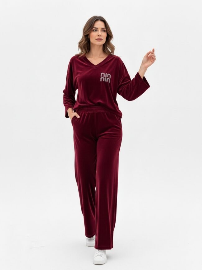 Woman in burgundy velvet loungewear set with a V-neck top and wide-leg pants, logo on the chest, walking forward in white sneakers against a white background.
