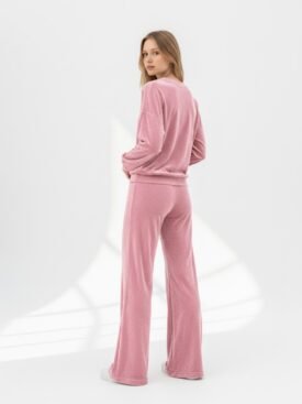 Model in a pink velour tracksuit facing away, standing in a bright studio with white background.