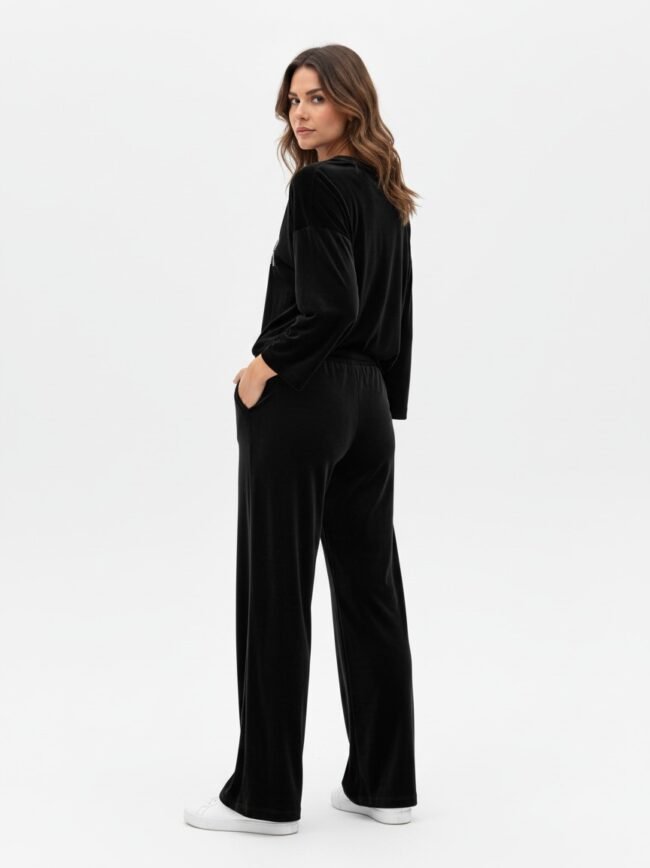 Woman in black velour loungewear: long-sleeve top and wide-leg pants, standing in profile with hands in pockets, white sneakers against a white studio backdrop.
