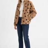 Woman wearing a tan teddy coat with black polka dots, a white turtleneck, dark jeans, and white sneakers, standing against a light background.