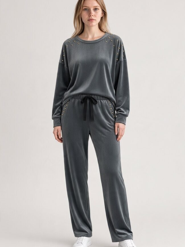 Velvet Γκρί Gray velour tracksuit with gold stud embellishments on shoulders and pockets, a black drawstring waist, and white sneakers on a standing model, fashion photo.