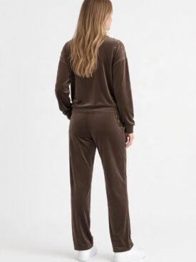 Woman wearing a brown velour tracksuit seen from behind, with gold studs on the shoulders and side, and white sneakers.