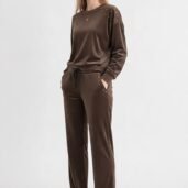Woman in a brown velour tracksuit with gold studs on shoulders and pockets, standing with hands in pockets, wearing white sneakers.