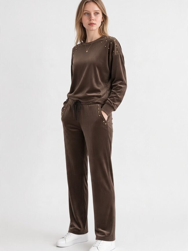 Velvet Καφέ 3 Woman in a brown velour tracksuit with gold studs on shoulders and pockets, standing with hands in pockets, wearing white sneakers.